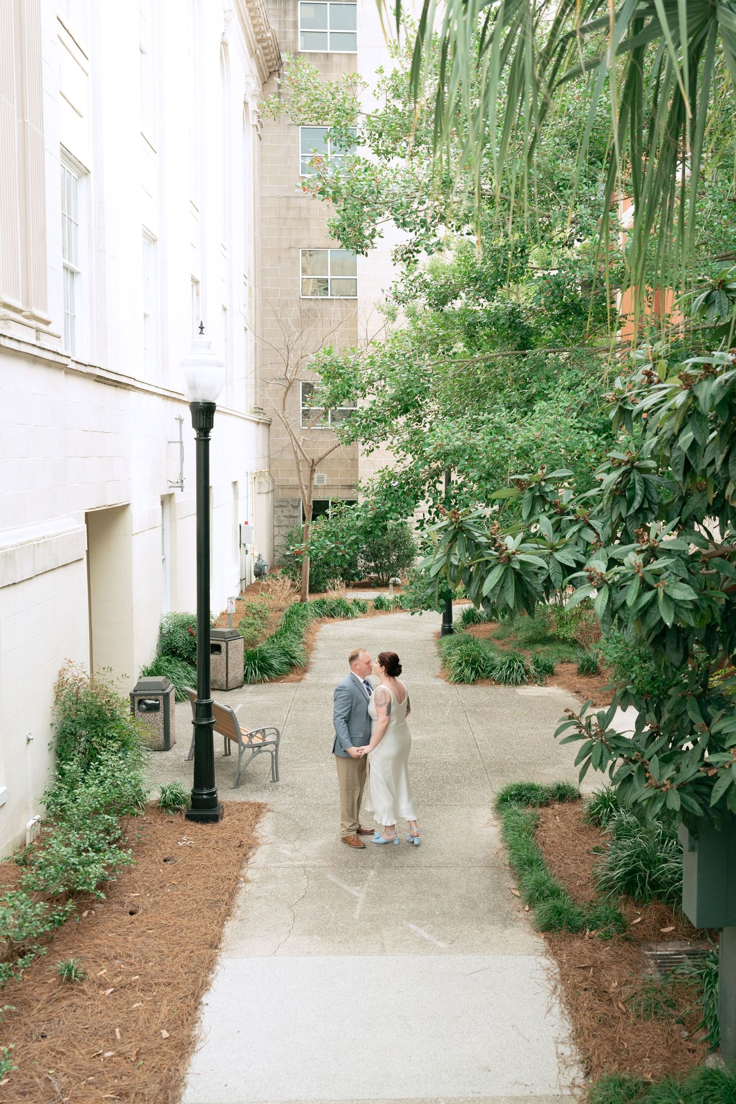 A couple stands close together on a peaceful garden path surrounded by lush greenery and brick buildings.