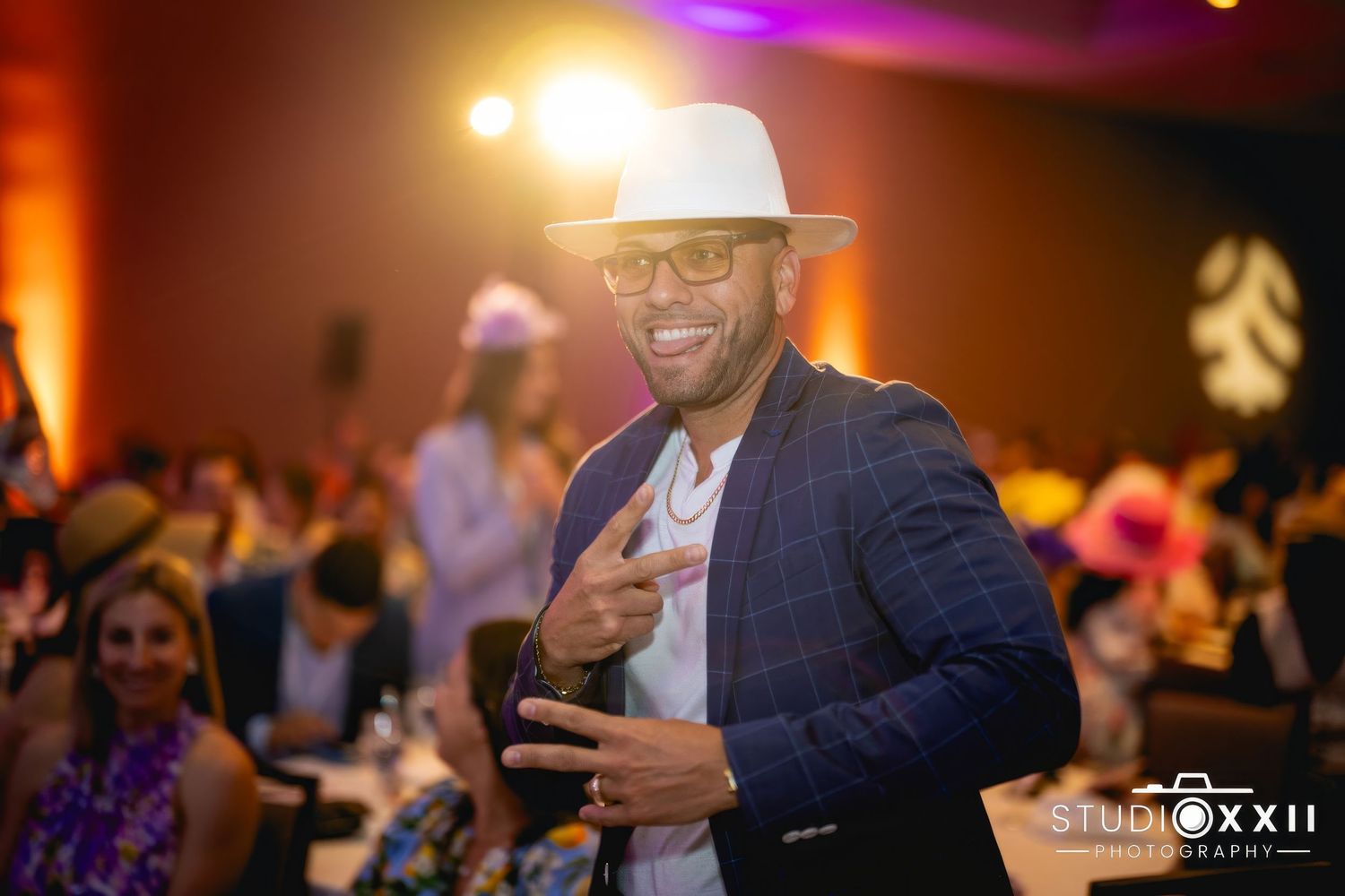 Smiling guest in a dark blazer and white fedora hat enjoying an evening celebration with colorful lighting.
