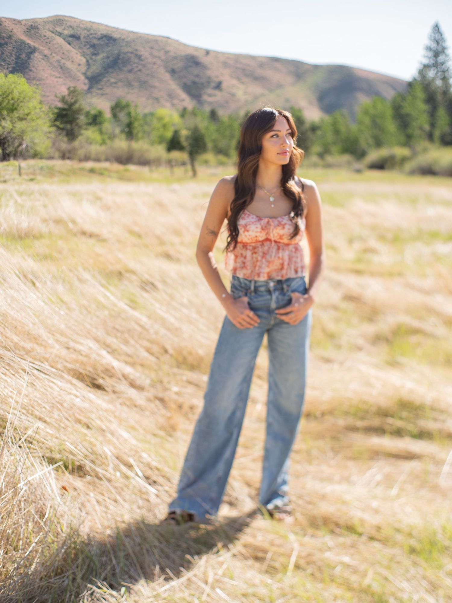 A high school senior stands in a golden field wearing a floral top and wide-leg jeans with mountains and trees in the background.