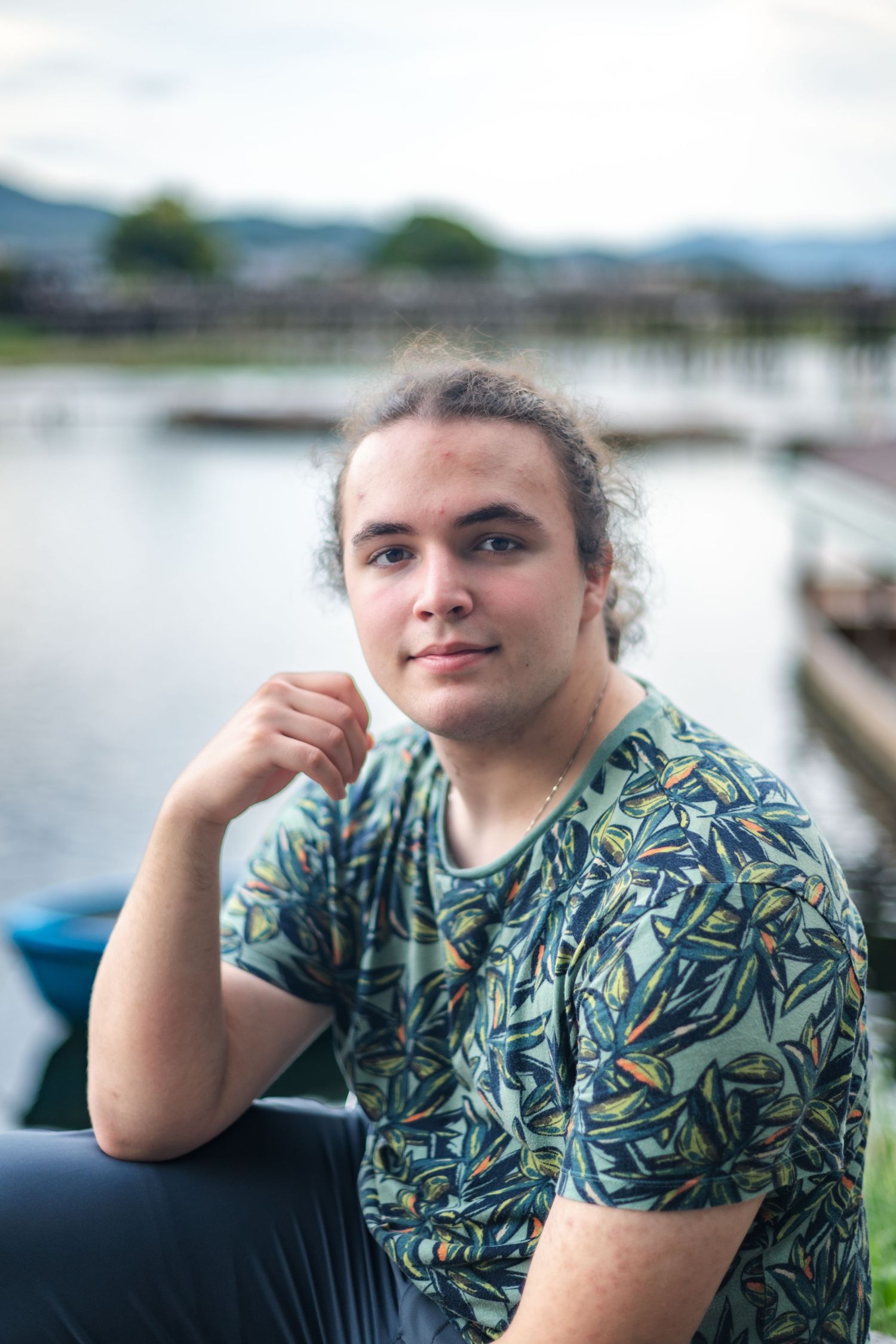 Someone wearing a tropical print shirt sits near a marina with boats and water in the background on an overcast day.