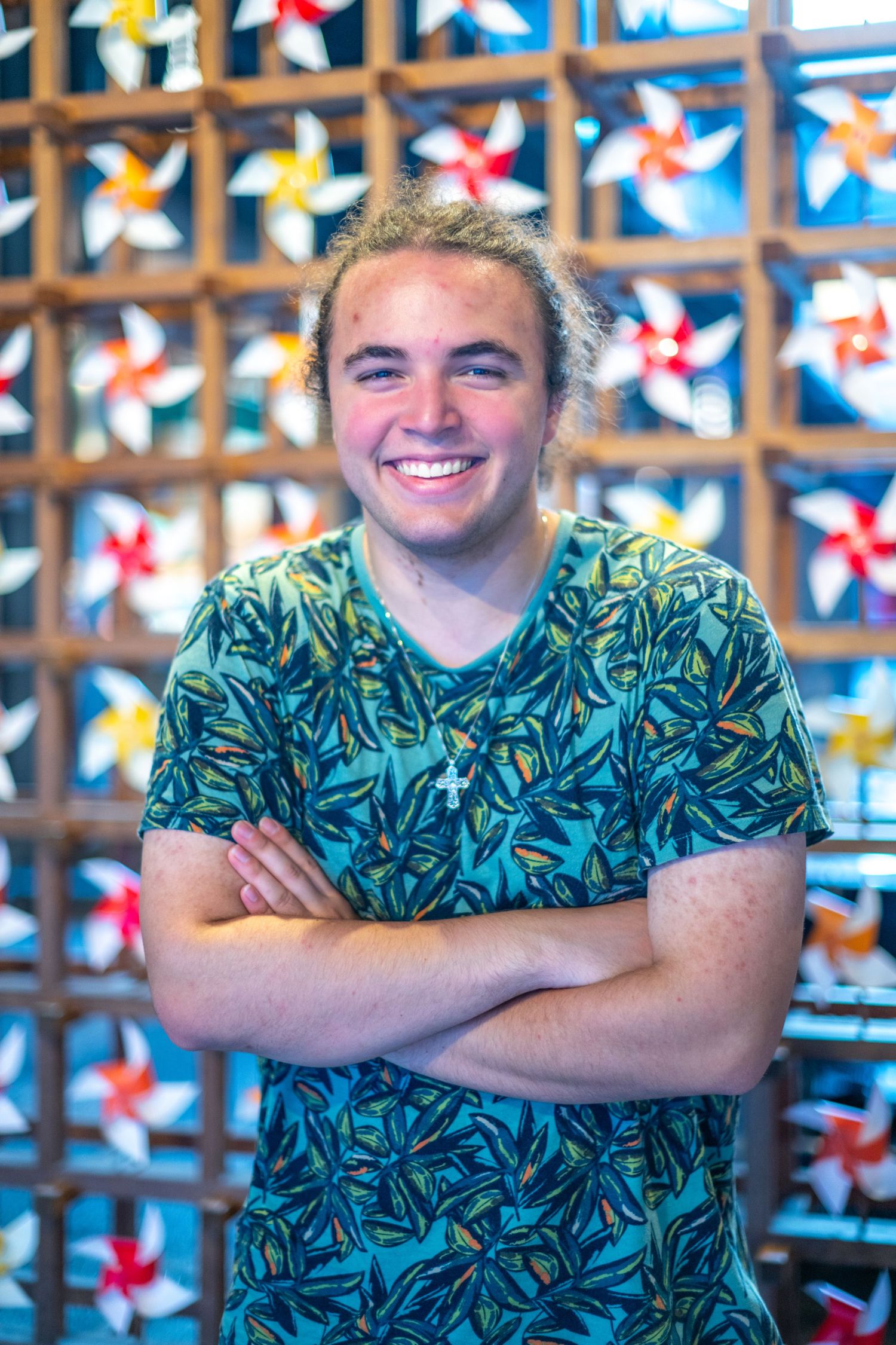 Hawaiian shirt-clad individual poses with crossed arms against colorful pinwheel wall display in bright lighting.
