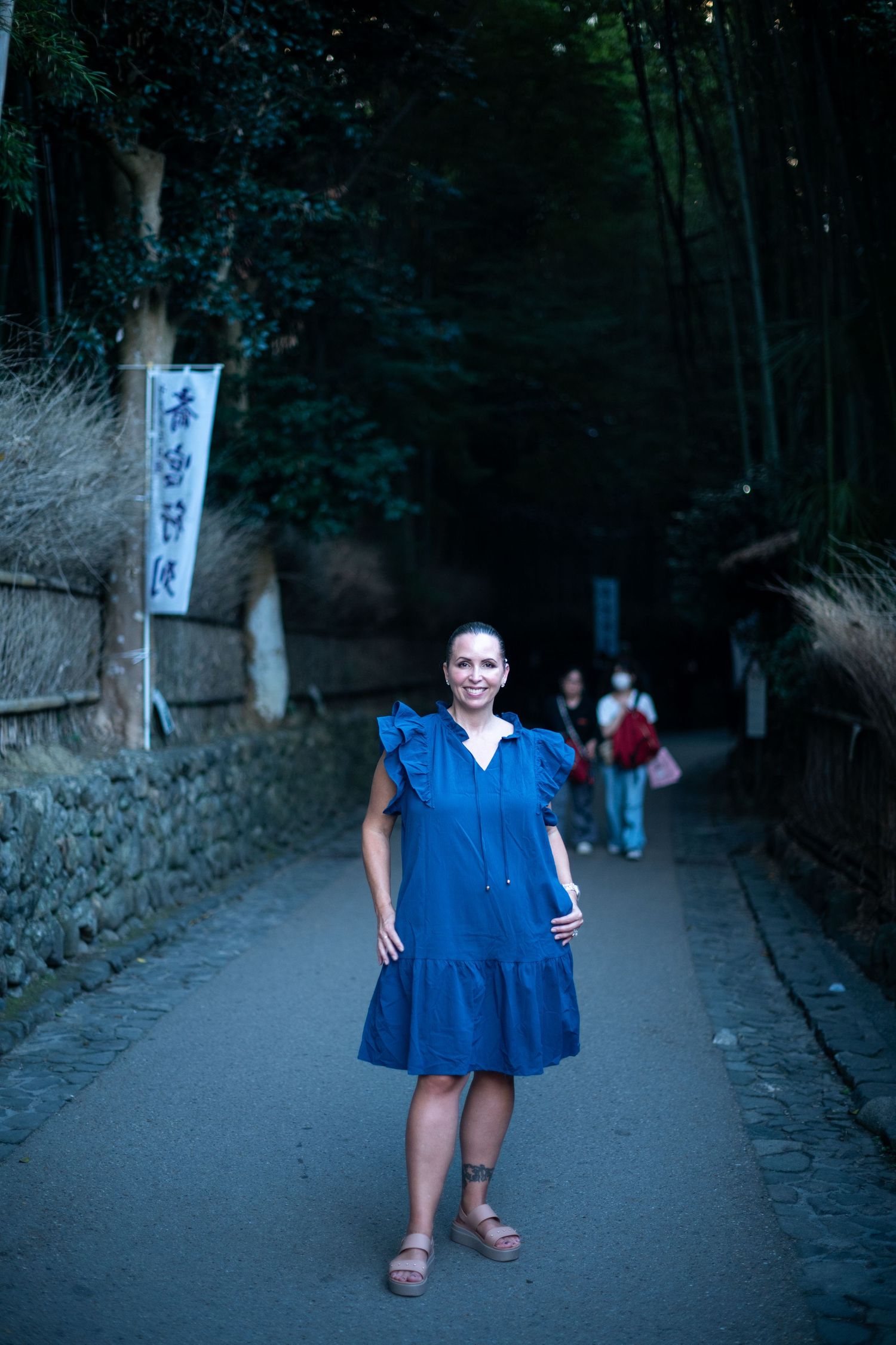 Someone wearing a blue dress poses on a shaded path in Japan with traditional stone walls and bamboo forest.