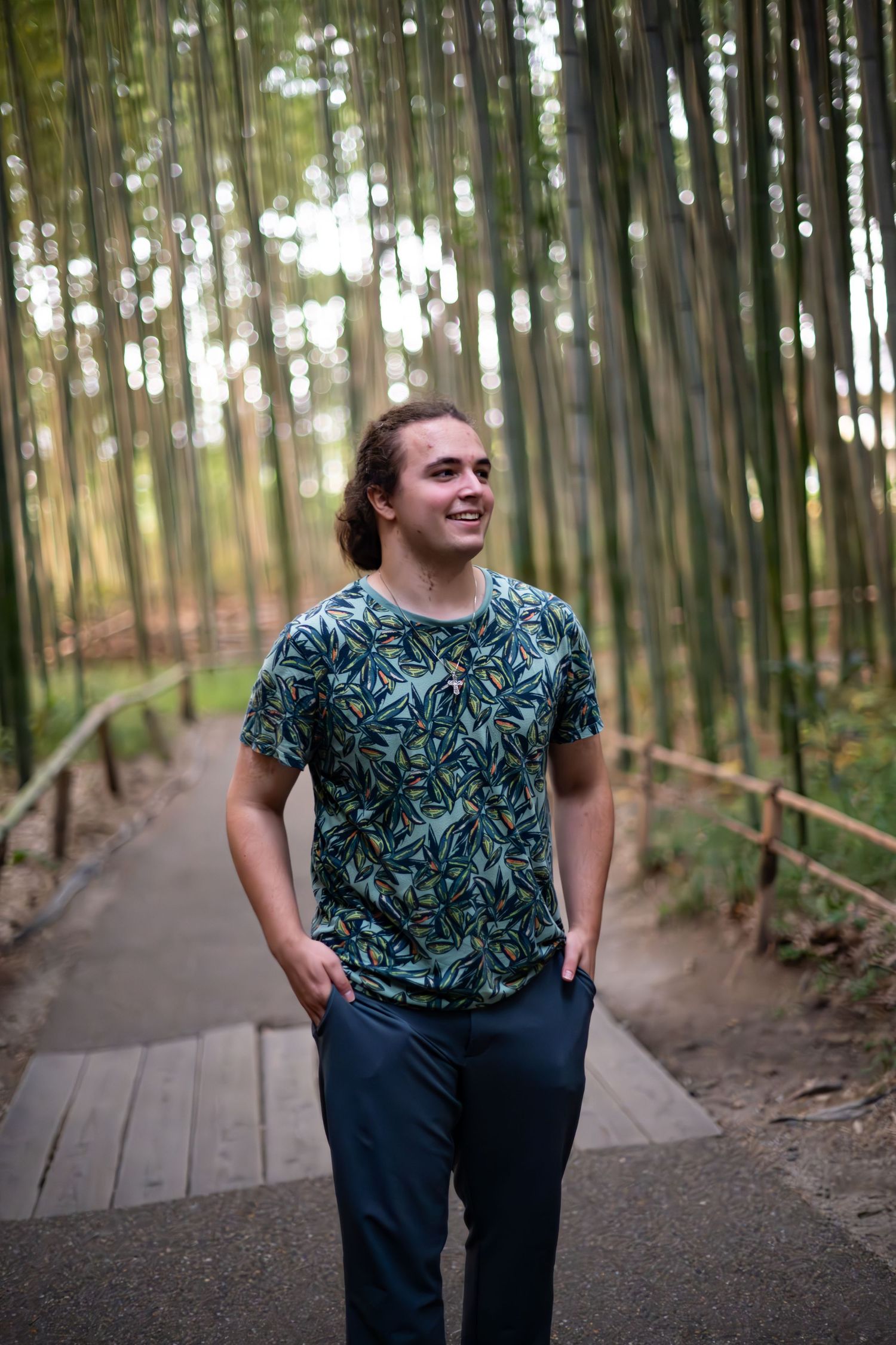 Person wearing a tropical print t-shirt smiles while walking along a wooden path through a serene bamboo forest.