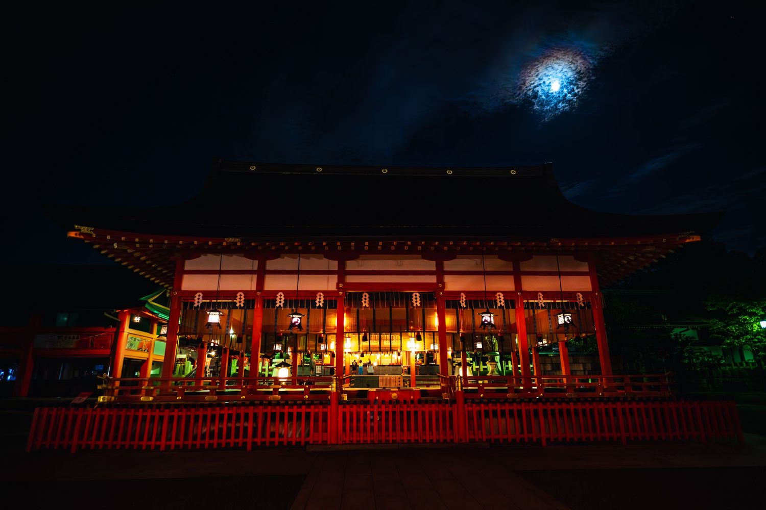 A traditional Japanese temple illuminated at night with red columns and a bright moon shining above through misty clouds.