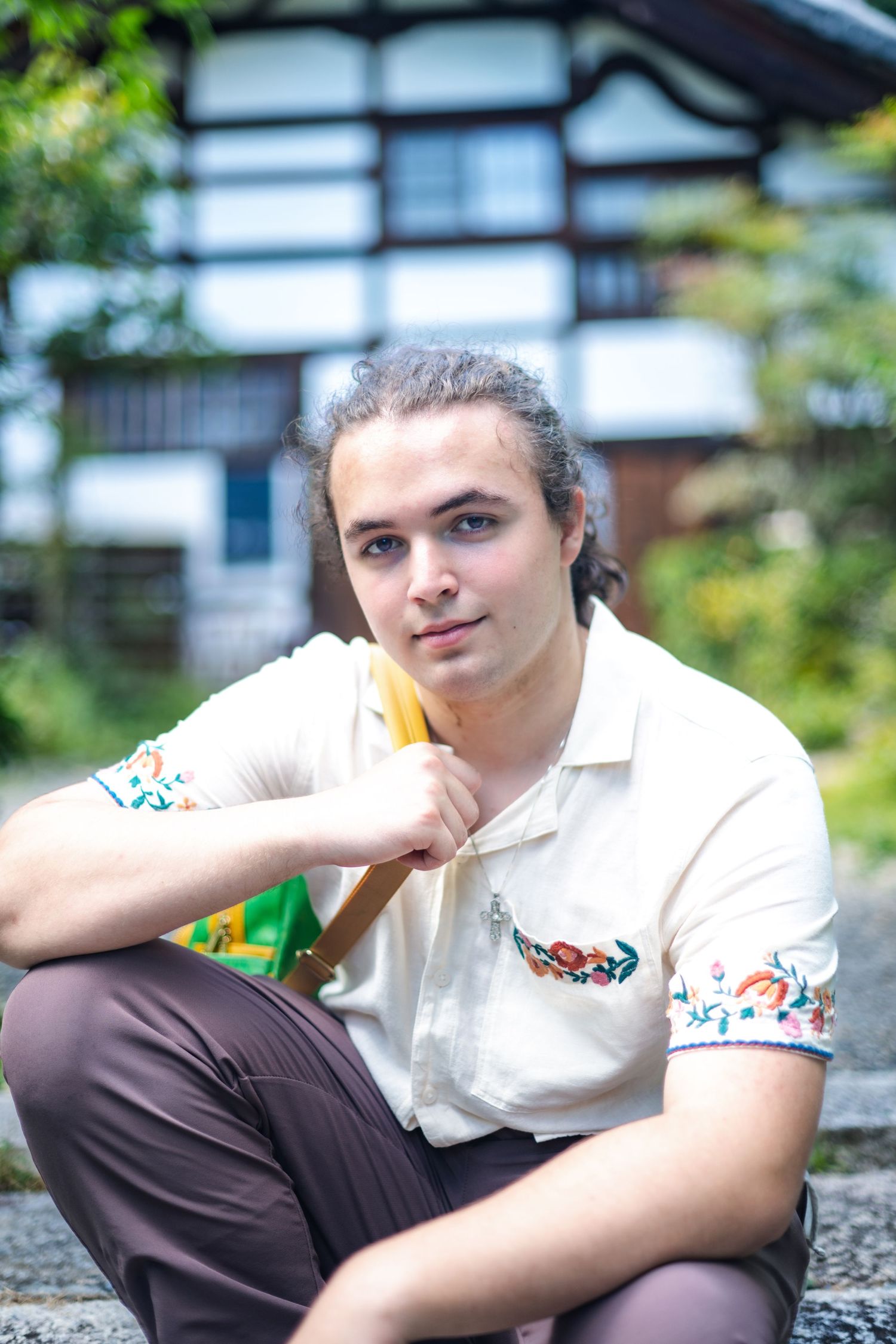 Someone wearing a white embroidered polo shirt sits casually in front of a traditional Japanese building in summer.