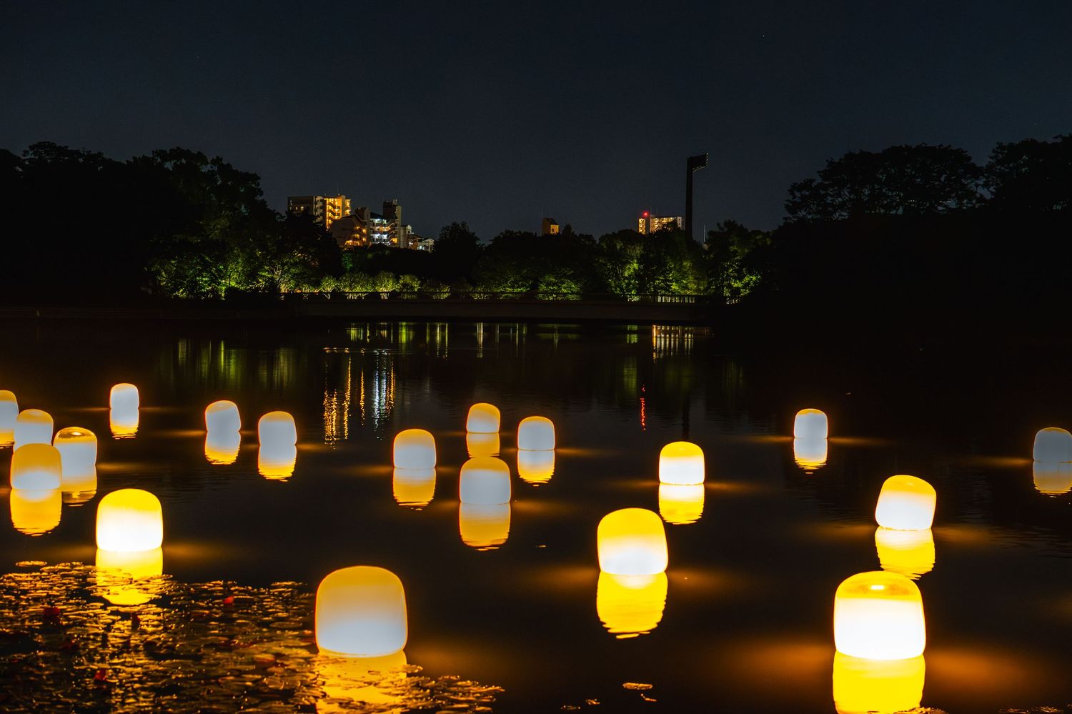 Glowing lanterns float on a dark lake at night with illuminated buildings and trees reflecting in the water.