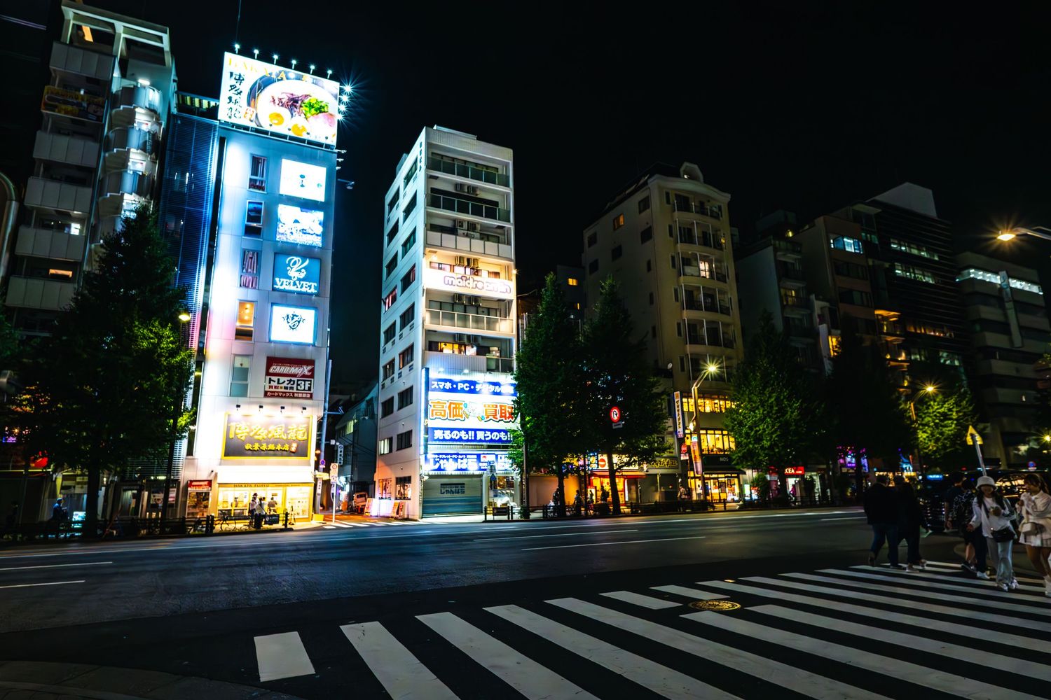 A nighttime street scene in Tokyo with illuminated buildings, neon signs, and a crosswalk on a busy urban corner.