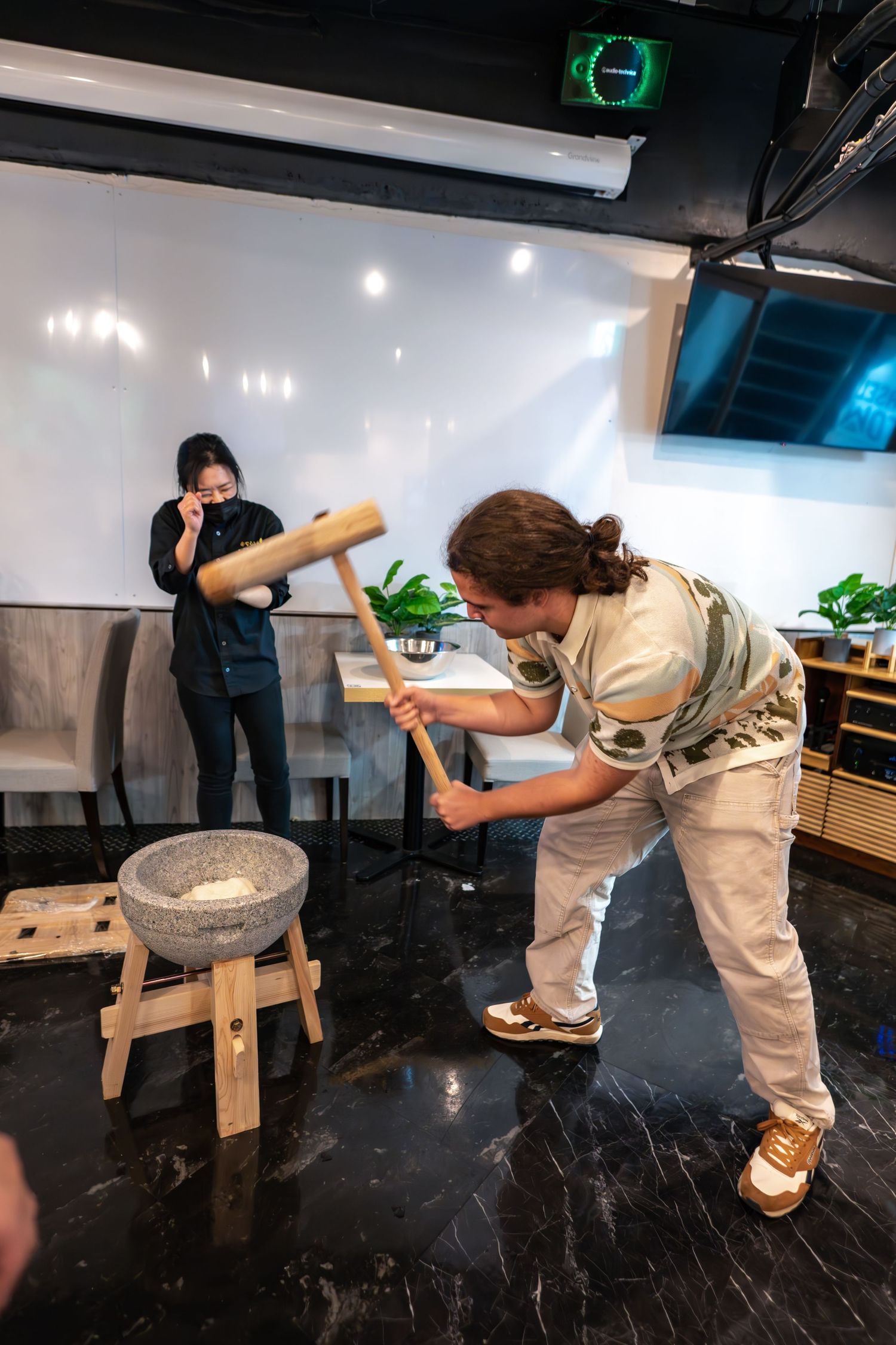 Two people demonstrate traditional cooking techniques with a stone grinder in a modern indoor space with plants and lighting.