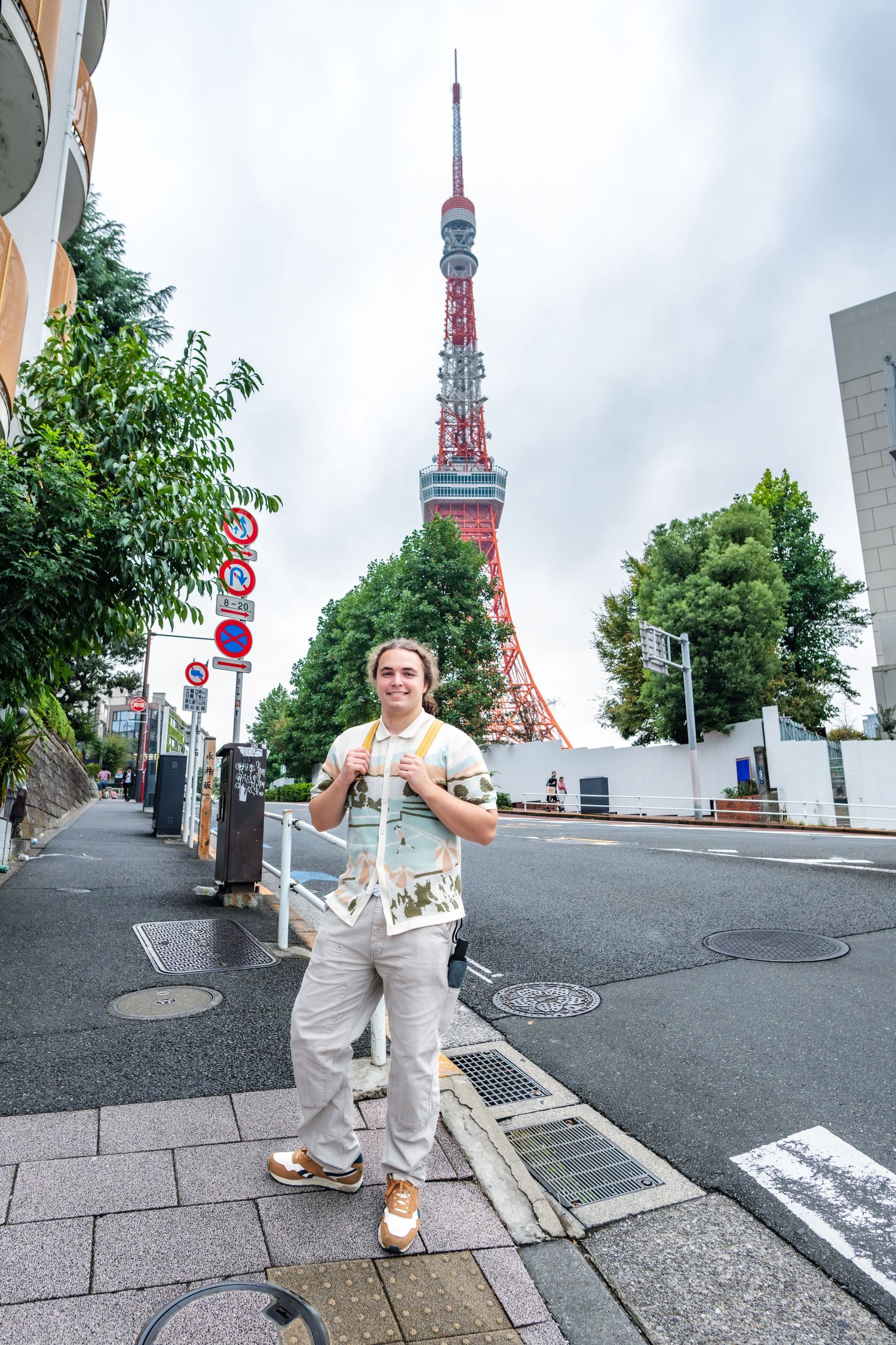Tourist taking photo on street with iconic Tokyo Tower rising in the background against cloudy sky.