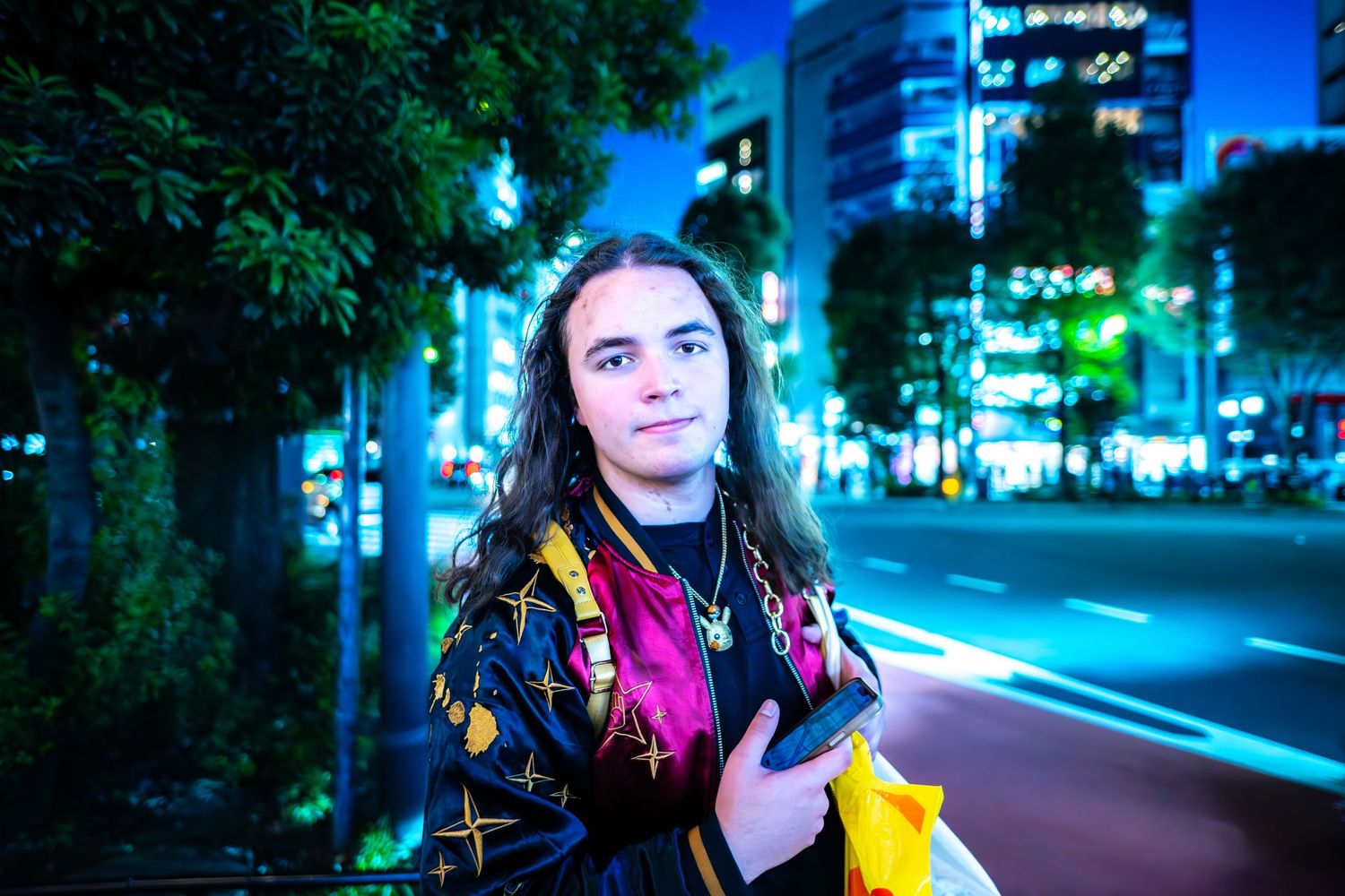 Nighttime street photo with colorful jacket against urban backdrop of glowing blue buildings and trees.