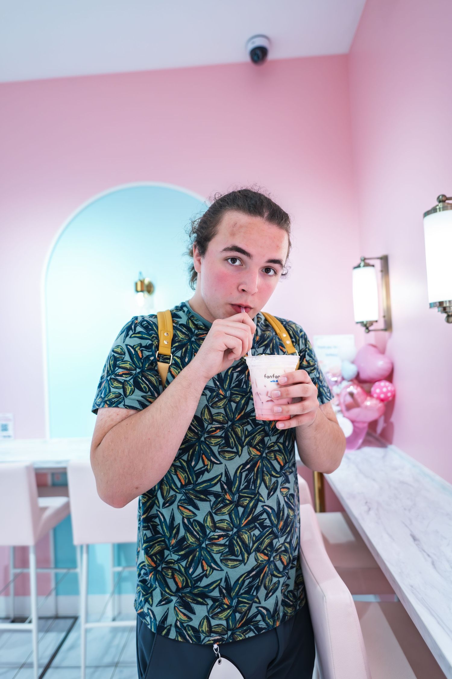 Someone in a tropical print shirt and suspenders drinks a beverage through a straw in a pink-walled cafe setting.