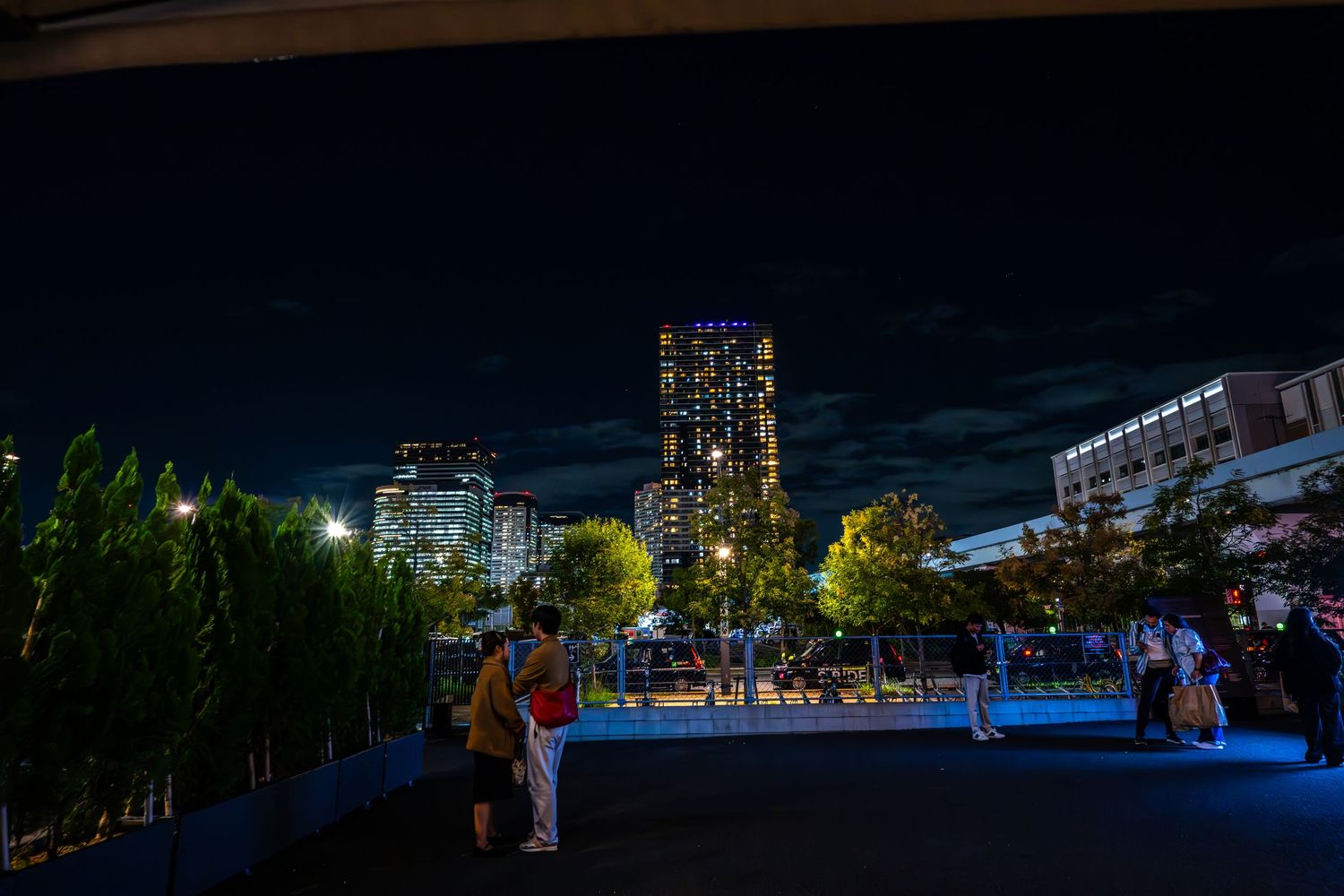 People gather on a rooftop terrace at night with illuminated city buildings and blue lighting creating an urban atmosphere.
