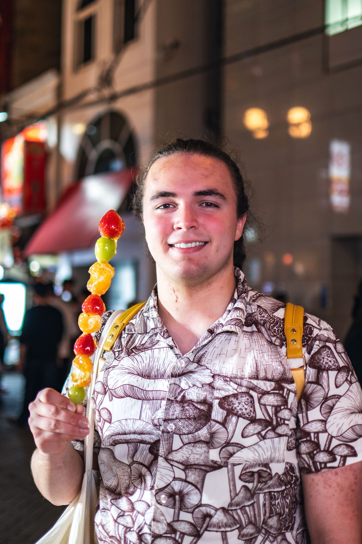 Person holding a colorful fruit skewer while wearing a patterned short-sleeve shirt on a city street at night.