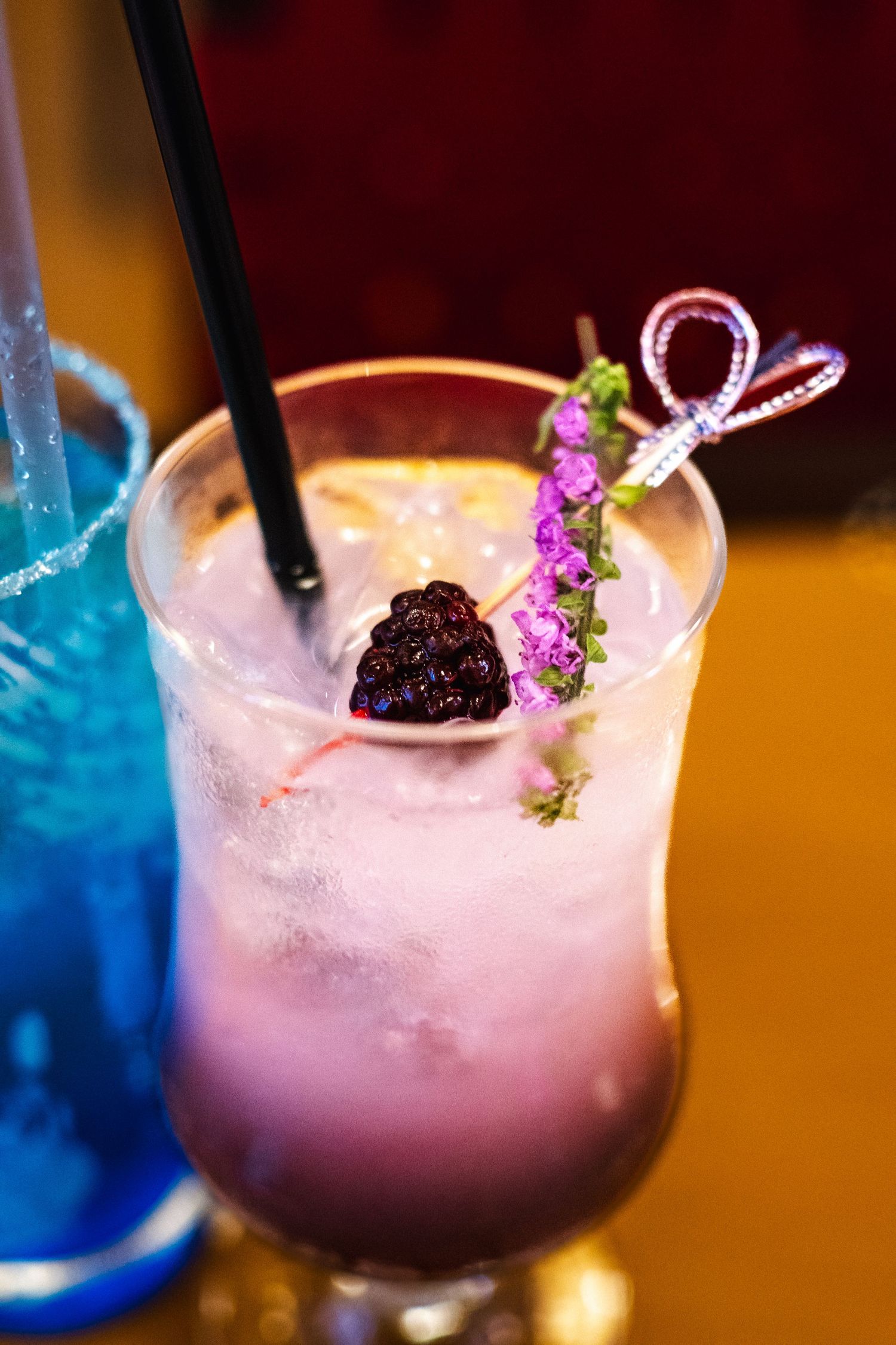 Pink cocktail garnished with fresh blackberry and purple flowers served in a tall glass with black straw and ice.