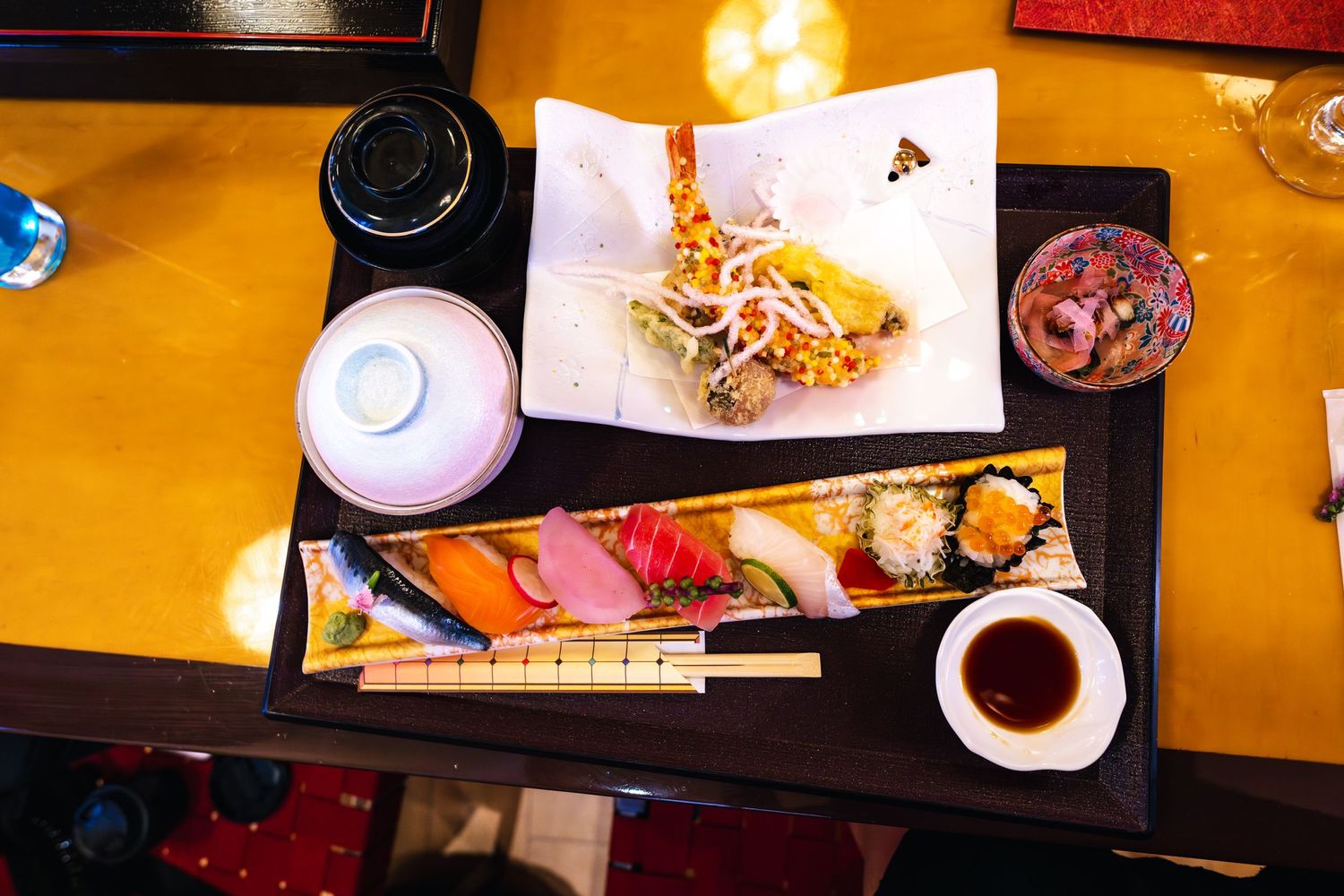 Overhead view of sushi platter with soy sauce and side dishes served on orange table surface.