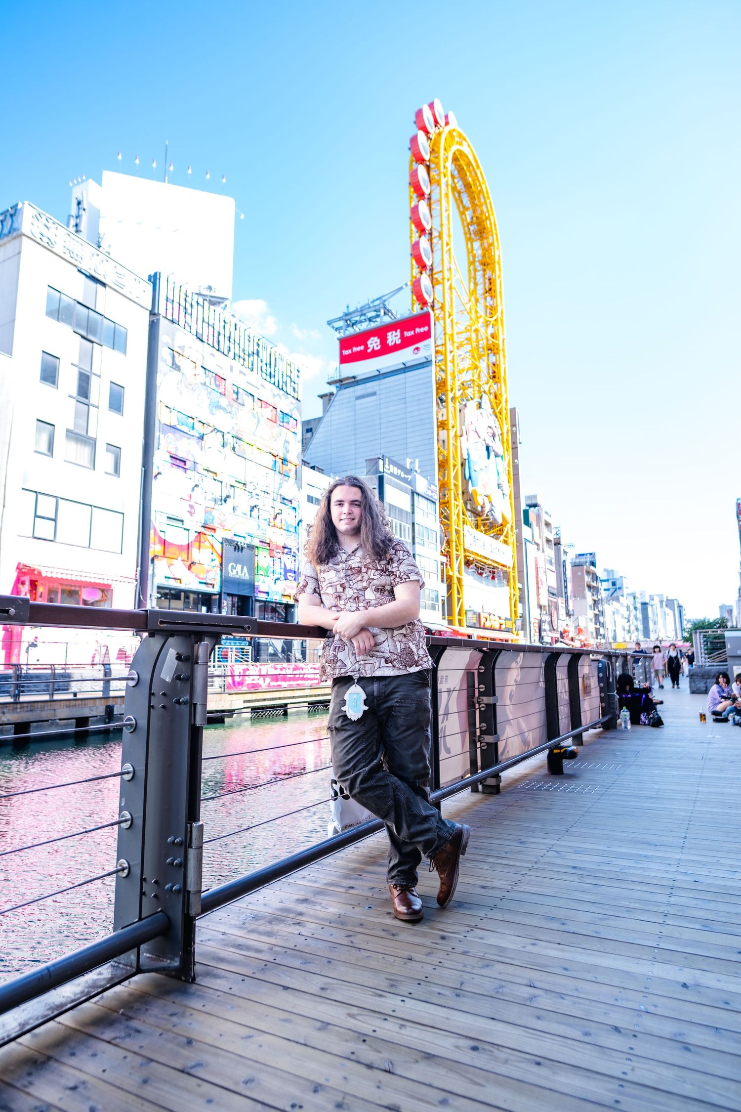 A boardwalk scene along the Dotonbori canal in Osaka with neon signs and billboards visible in the background.