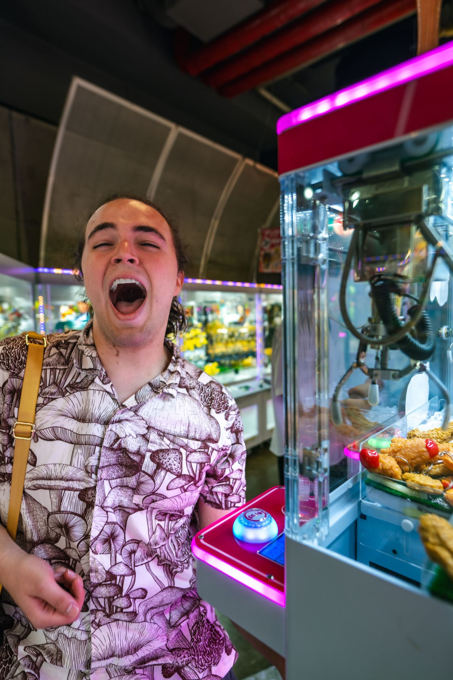 Person in patterned shirt laughs excitedly while standing next to an arcade claw machine game.