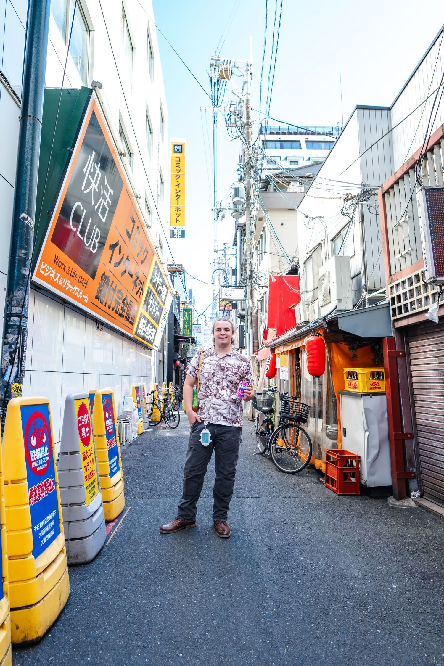 Narrow urban alley in Japan with yellow barriers, storefront signs, and power lines overhead on a sunny day.
