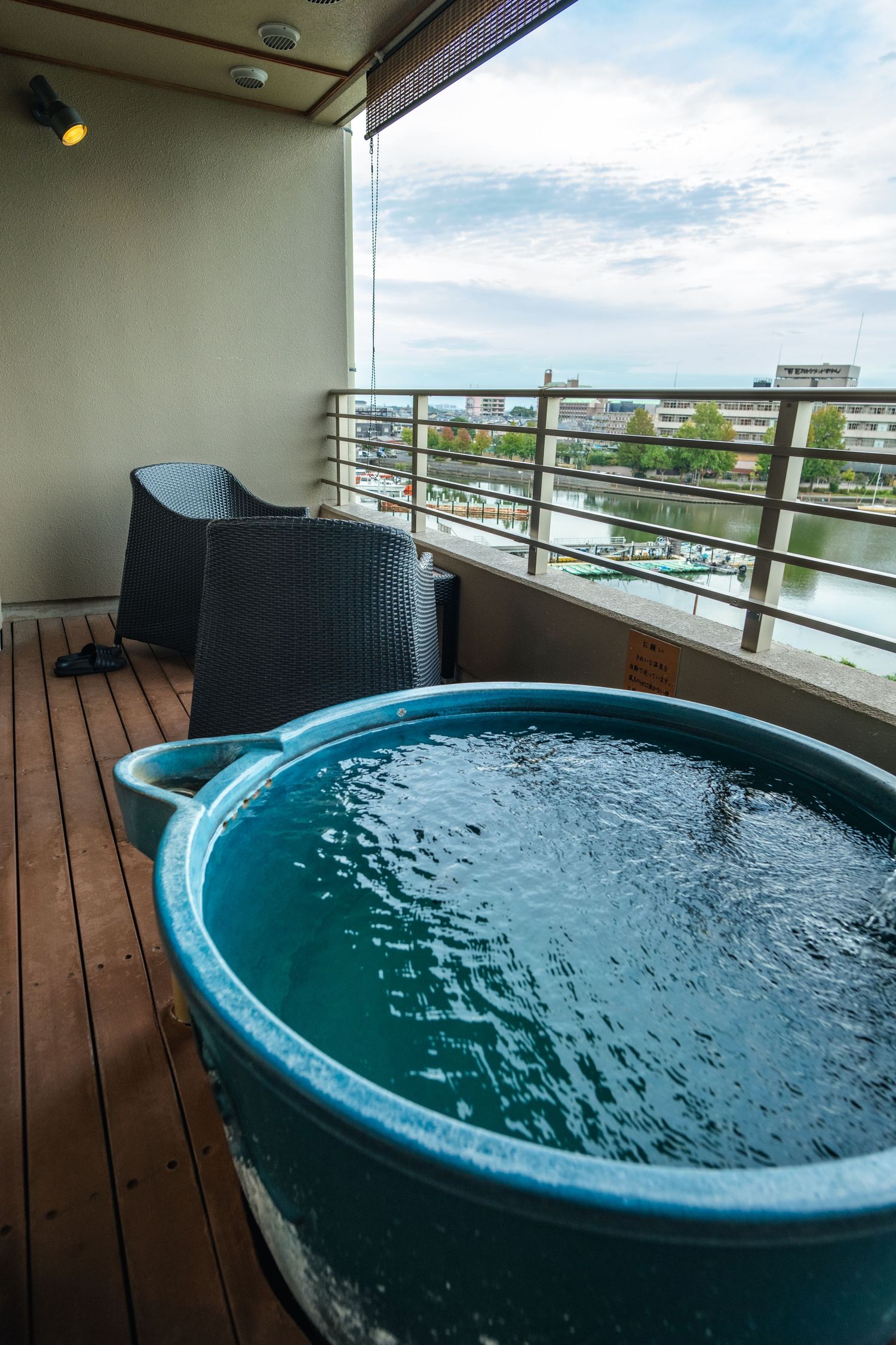 A round blue hot tub on a wooden balcony deck with outdoor furniture and city views in the background.