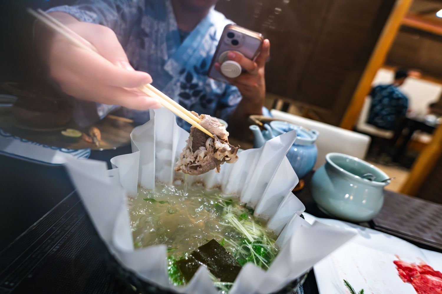 Someone adding fresh seafood and greens to a traditional Asian hotpot meal setup at a restaurant table.