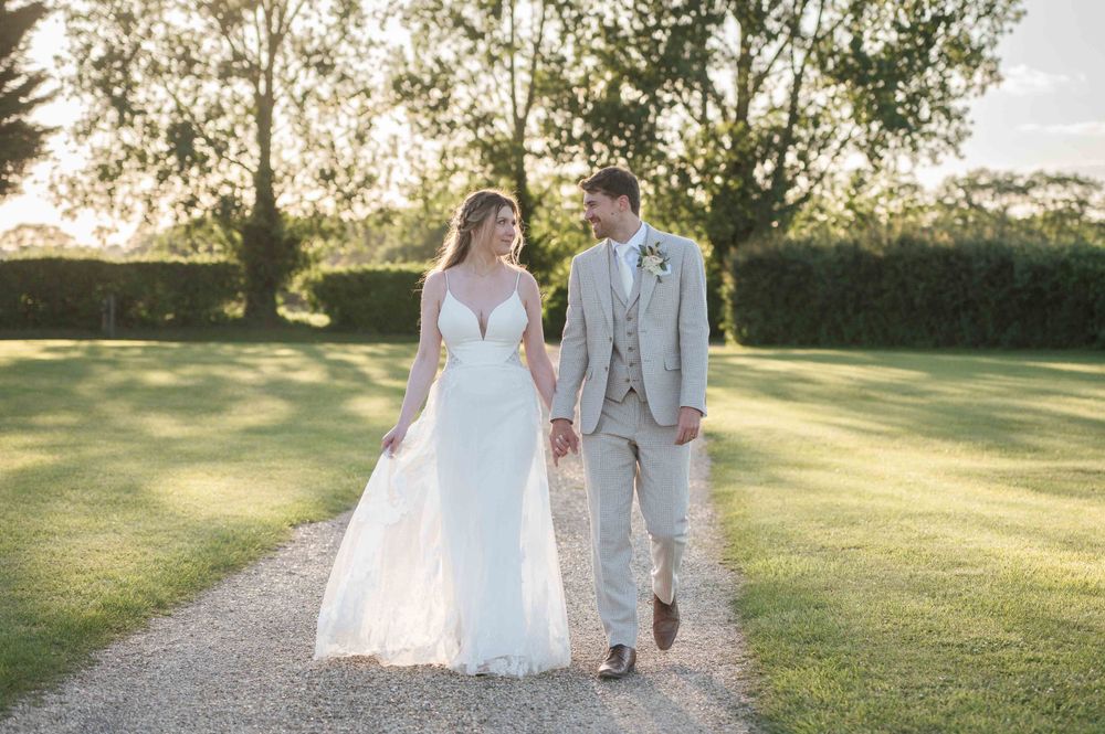 A couple walks hand in hand on a sunlit garden path, with the bride wearing a flowing white gown and groom in light gray suit.
