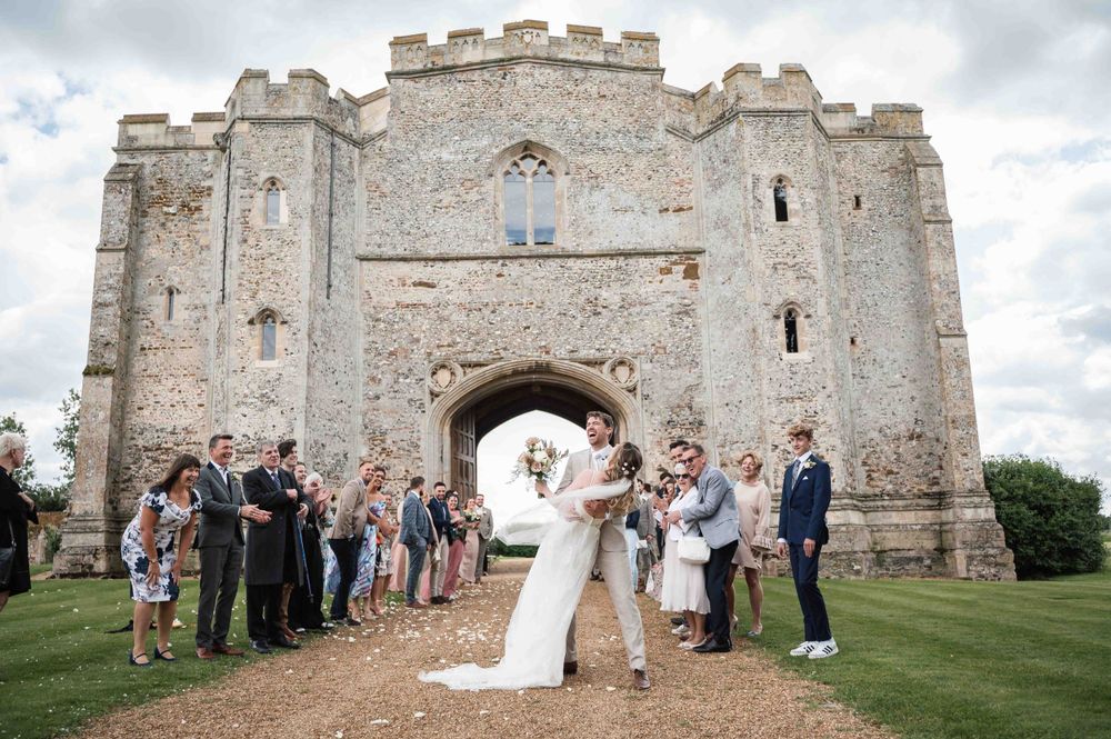 A wedding celebration taking place in front of a grand medieval stone castle entrance with guests gathered on the lawn.