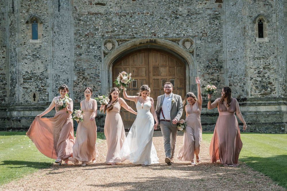 A wedding party walks together and laughs on a pathway in front of a historic stone castle on a sunny day.