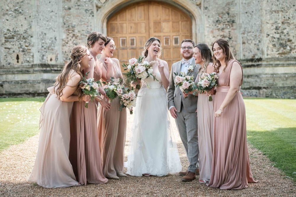 Wedding party wearing blush pink dresses and grey suits pose together holding floral bouquets outside stone church.