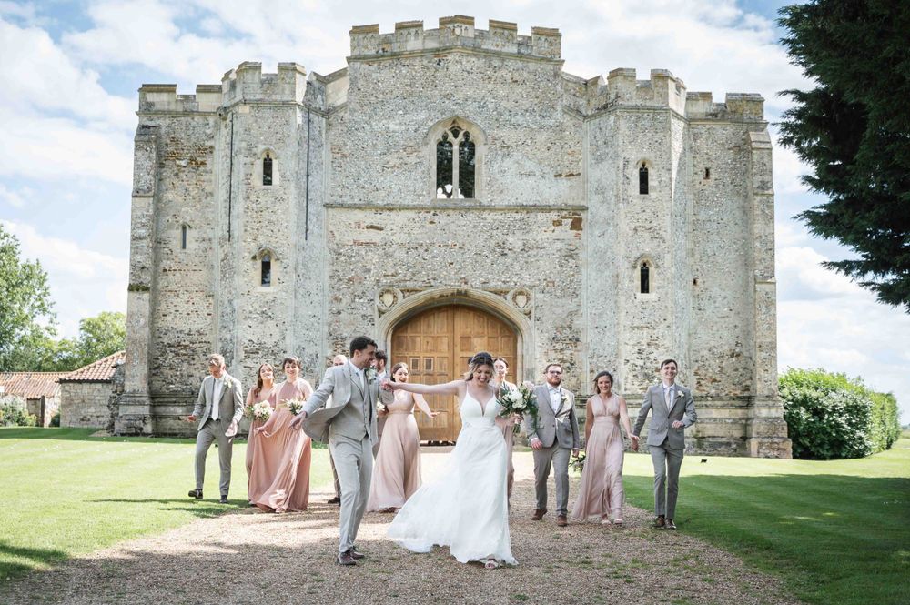 Wedding party in light colored attire poses at entrance of historic stone castle with crenellated towers.