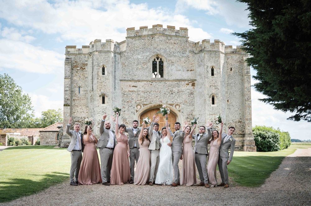 A wedding party poses together in front of a historic stone castle gatehouse with crenellated towers on a sunny day.
