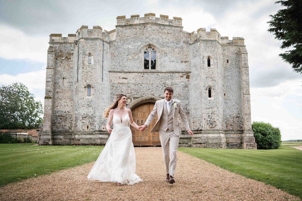 Couple in wedding attire walking hand in hand in front of a majestic stone castle with crenelated towers on a cloudy day.
