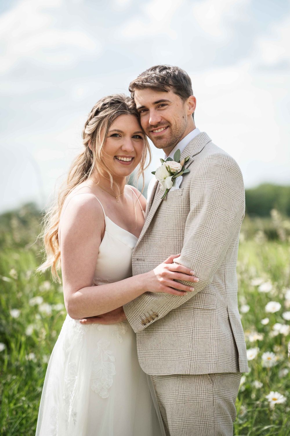 A couple embraces in a sunny meadow of daisies on their wedding day wearing light-colored formal attire.