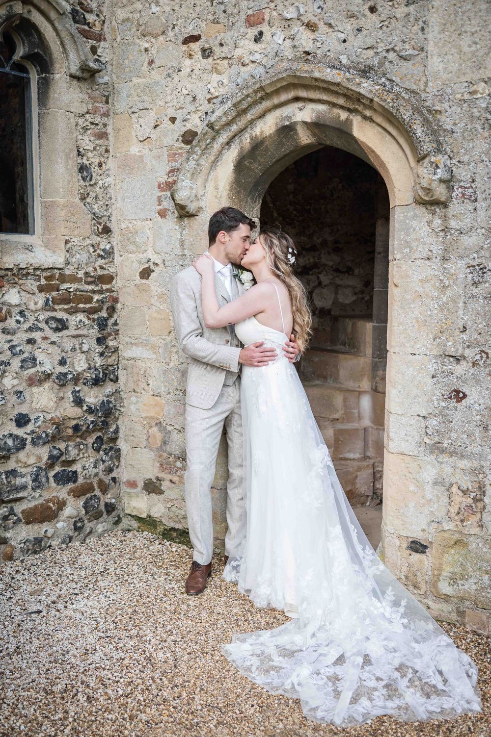 A couple embraces in an archway of an old stone church with a long lace wedding dress train trailing behind.