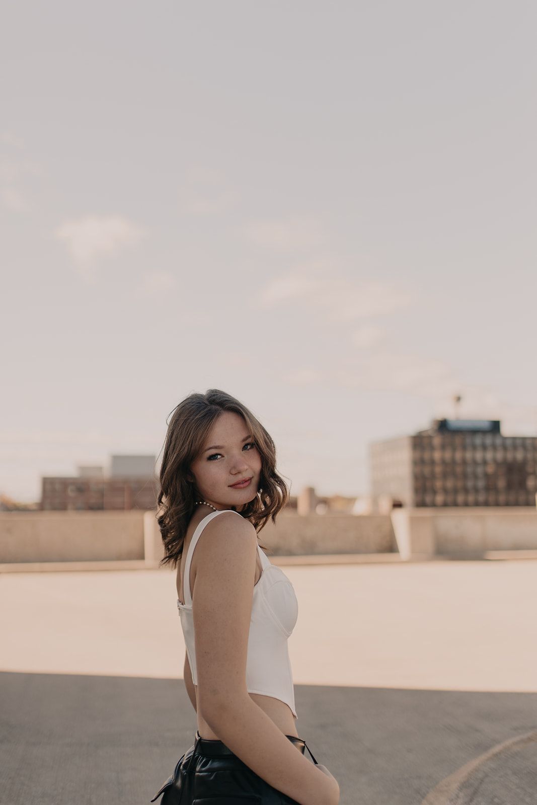A woman in a white tank top poses on an urban rooftop during golden hour.