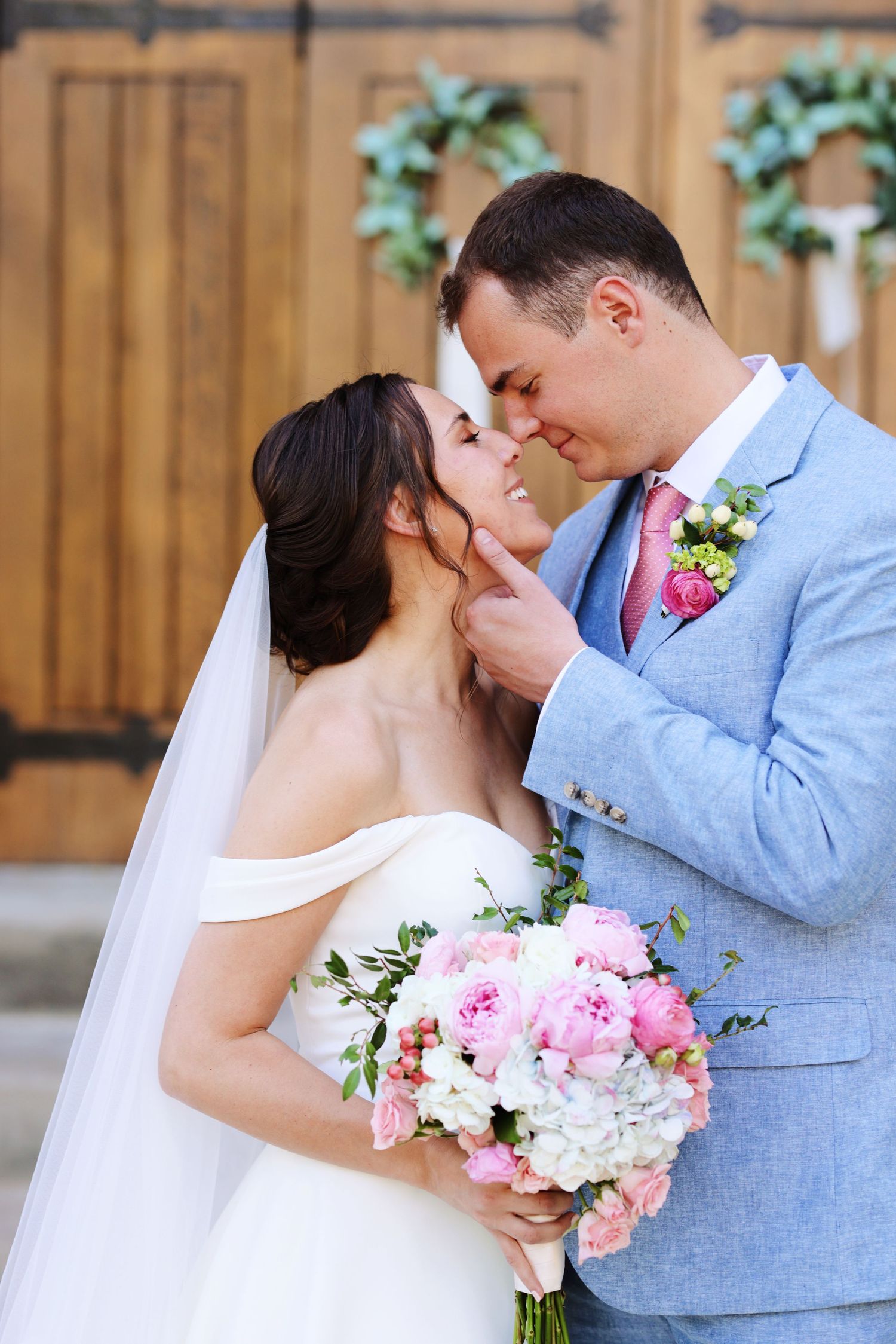 Romantic wedding portrait with couple embracing outdoors near wooden doors with floral decorations.
