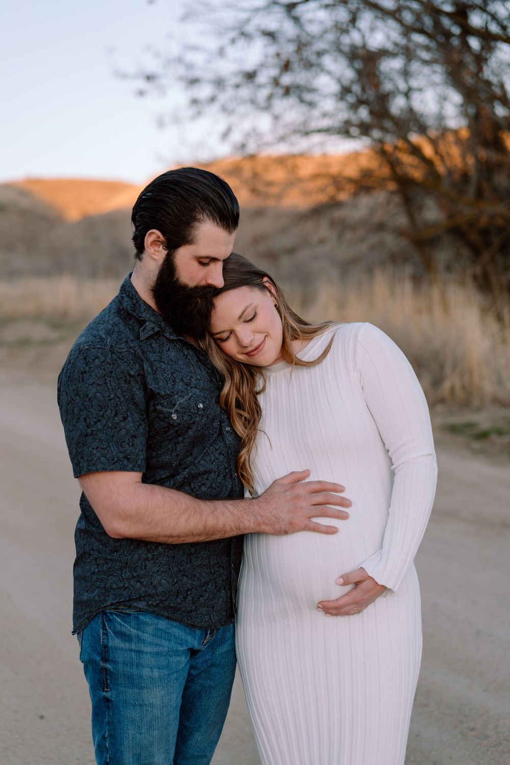 Expecting couple in casual attire embrace outdoors during sunset in natural setting with warm lighting.