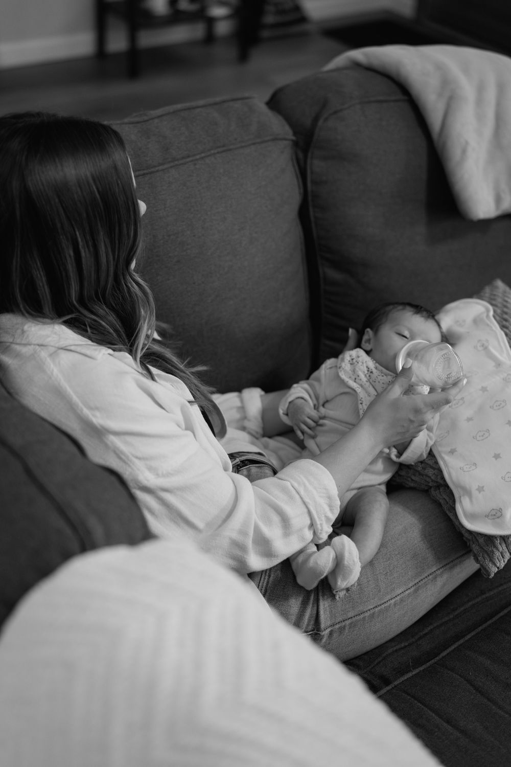 A tender black and white moment captured on a couch as a parent feeds a newborn baby with a bottle.