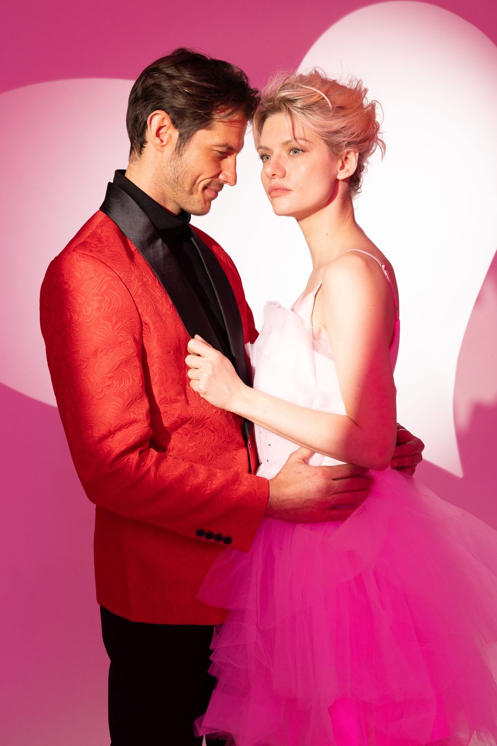 Romantic models embracing in formal wear against pink heart backdrop.