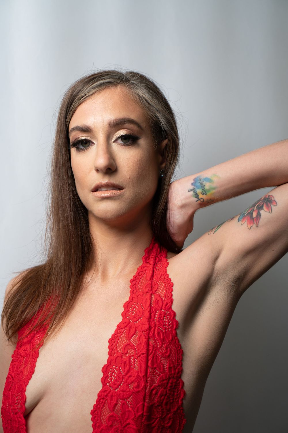 Boudoir image of a woman in red lace top shows colorful tattoos on arm against light background.