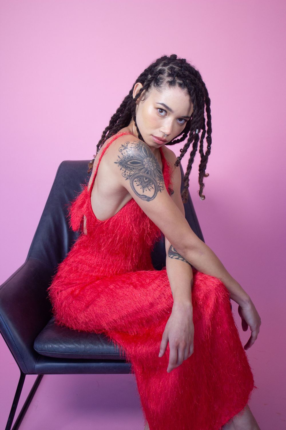 Model in red textured dress poses on chair against pink background, showing arm tattoo.