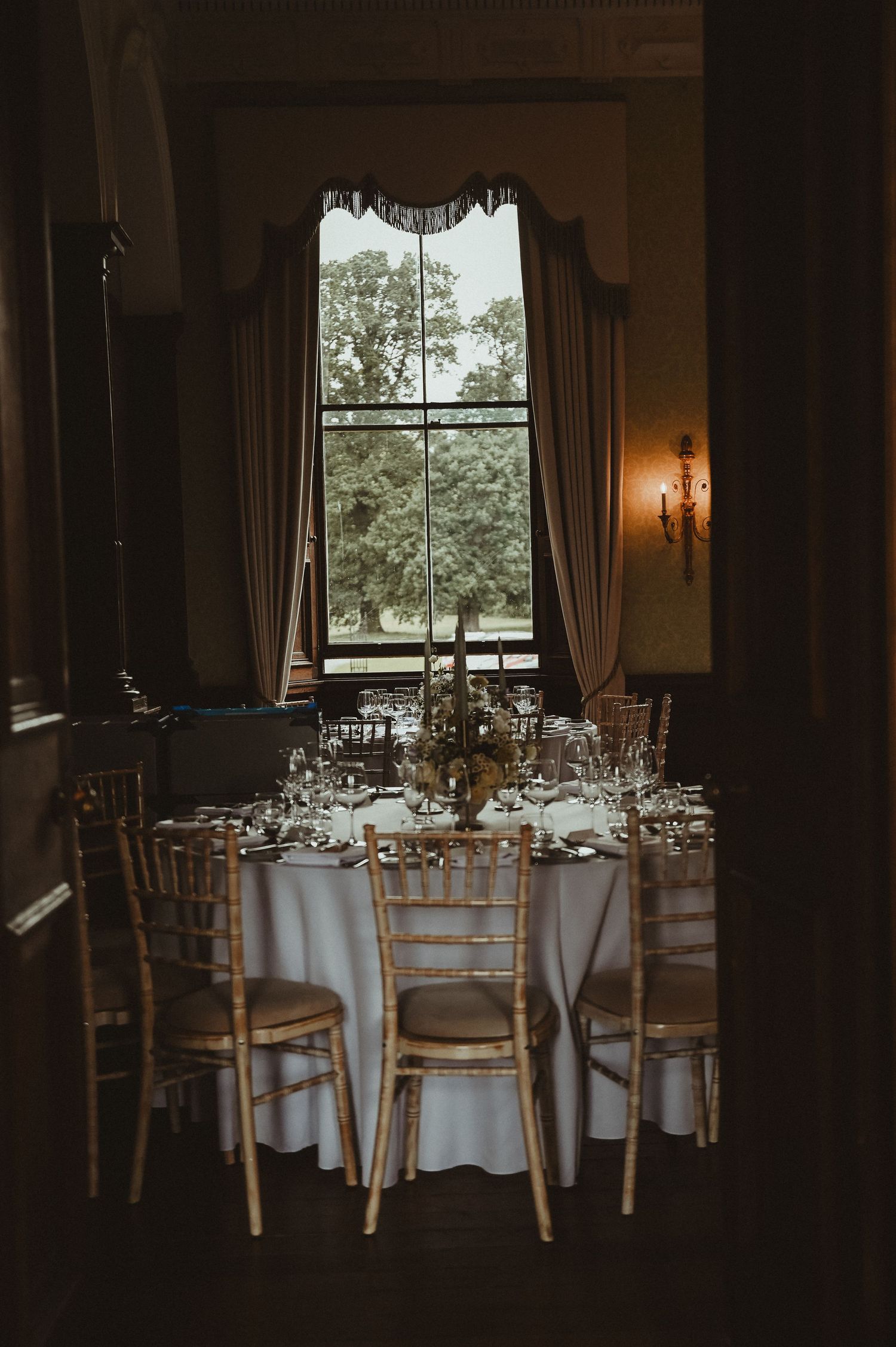 Elegant wedding reception table with white linens and gold chiavari chairs set against a tall window with draped curtains.