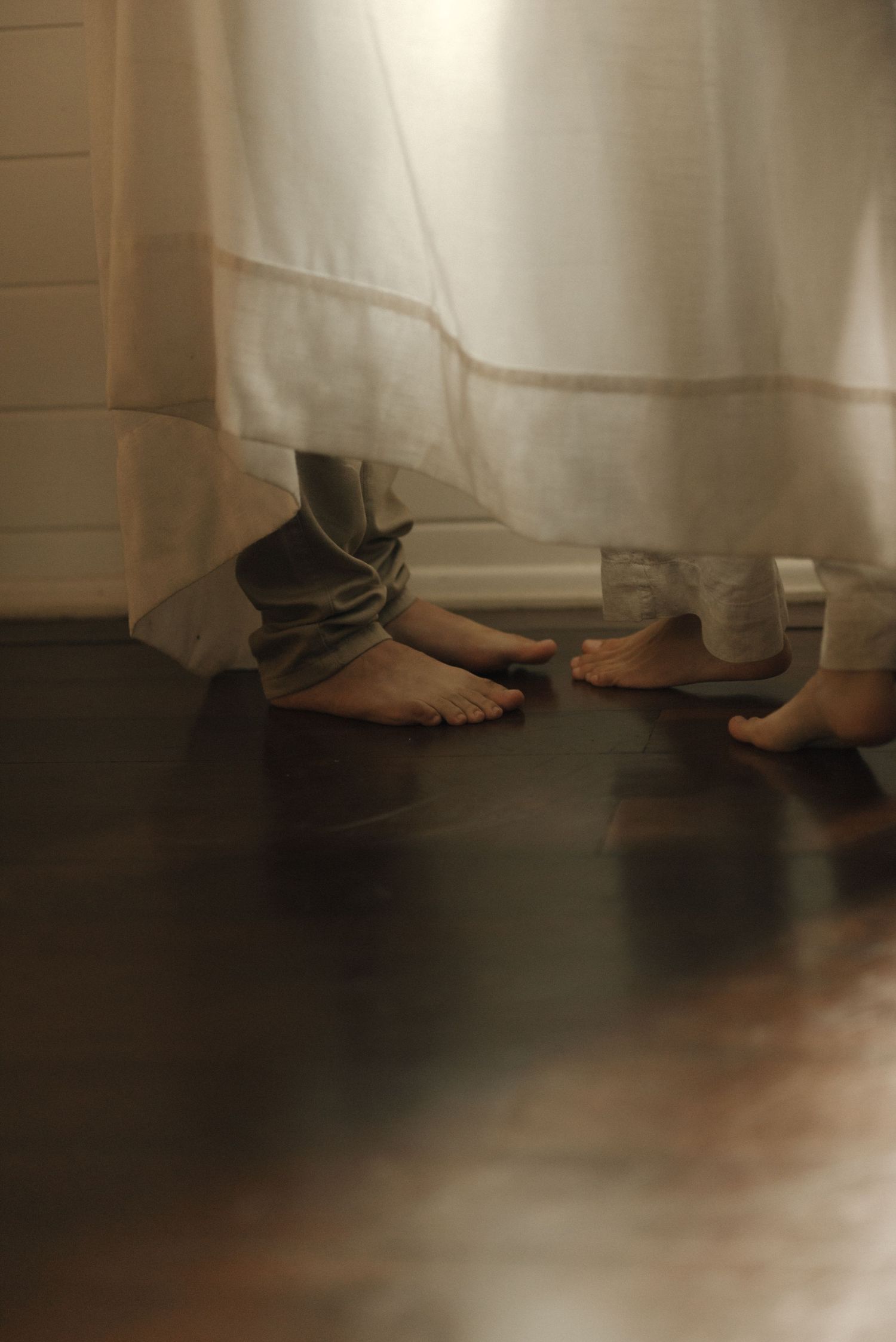 Bare feet peeking out from under white curtains on a dark wooden floor in moody lighting.