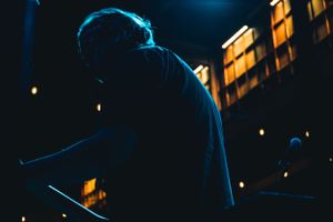 A silhouetted performer on stage illuminated by blue lighting with glowing windows visible in the background.