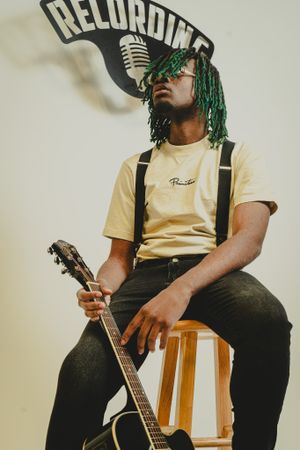 A musician with green dreadlocks sits on a stool holding a guitar under a recording sign in a vintage-style photo setup.