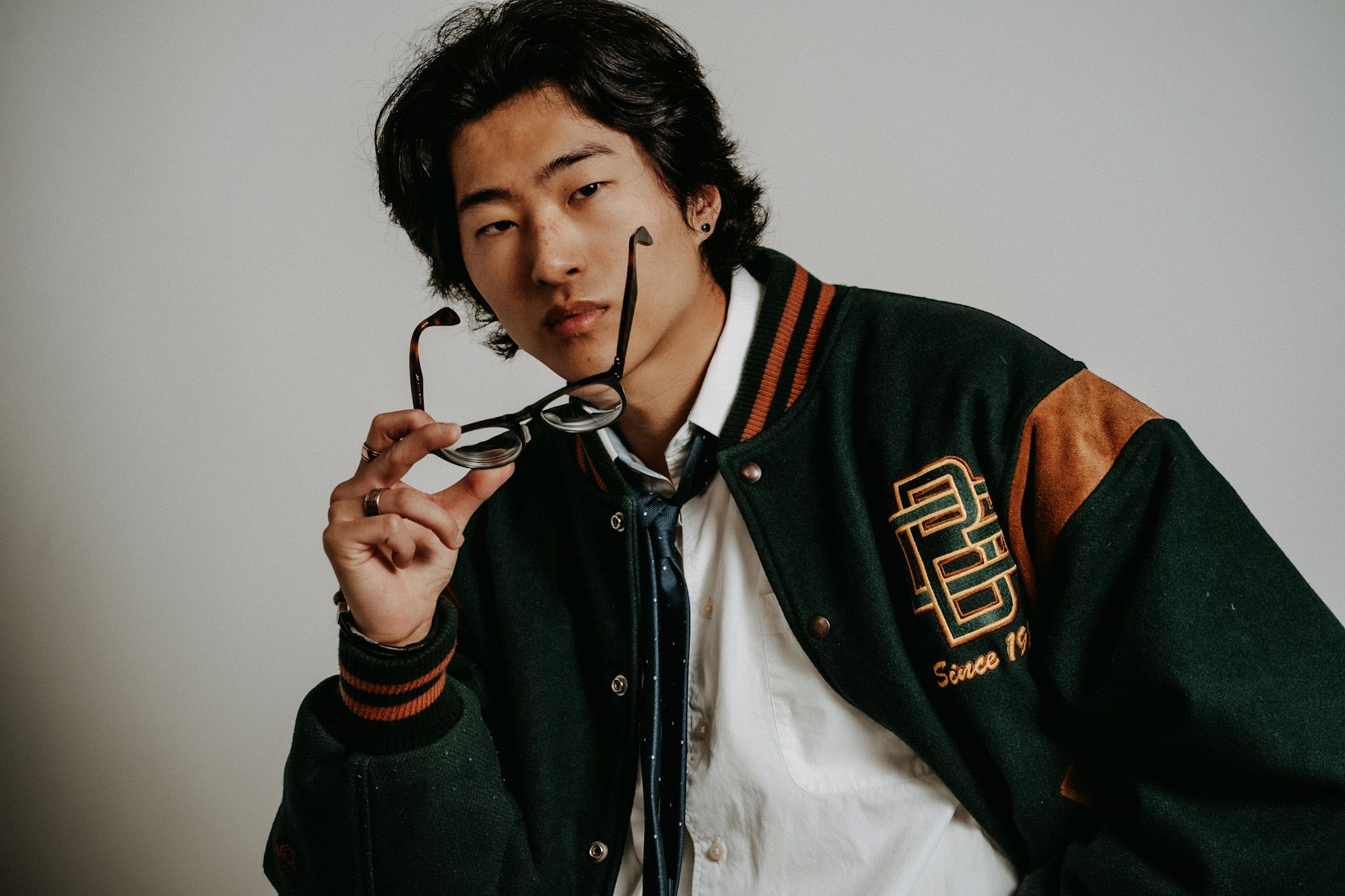 Studio portrait of a person in a varsity jacket holding glasses against a white background.