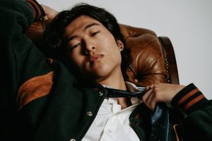 Person lounging on brown leather chair wearing dark jacket and white shirt in moody lighting.