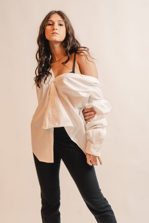 Fashion model poses in white off-shoulder button-up shirt and dark pants against light background.