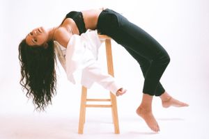 Model in white top and black pants arches backwards over wooden stool in studio.