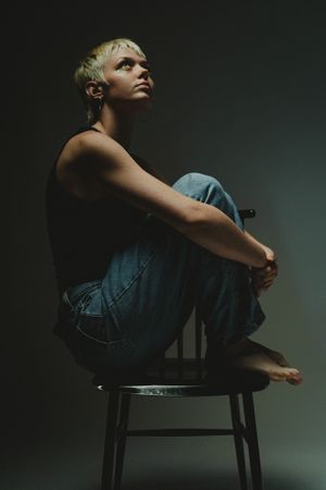 Model in denim sits curled up on wooden chair against dark studio backdrop.