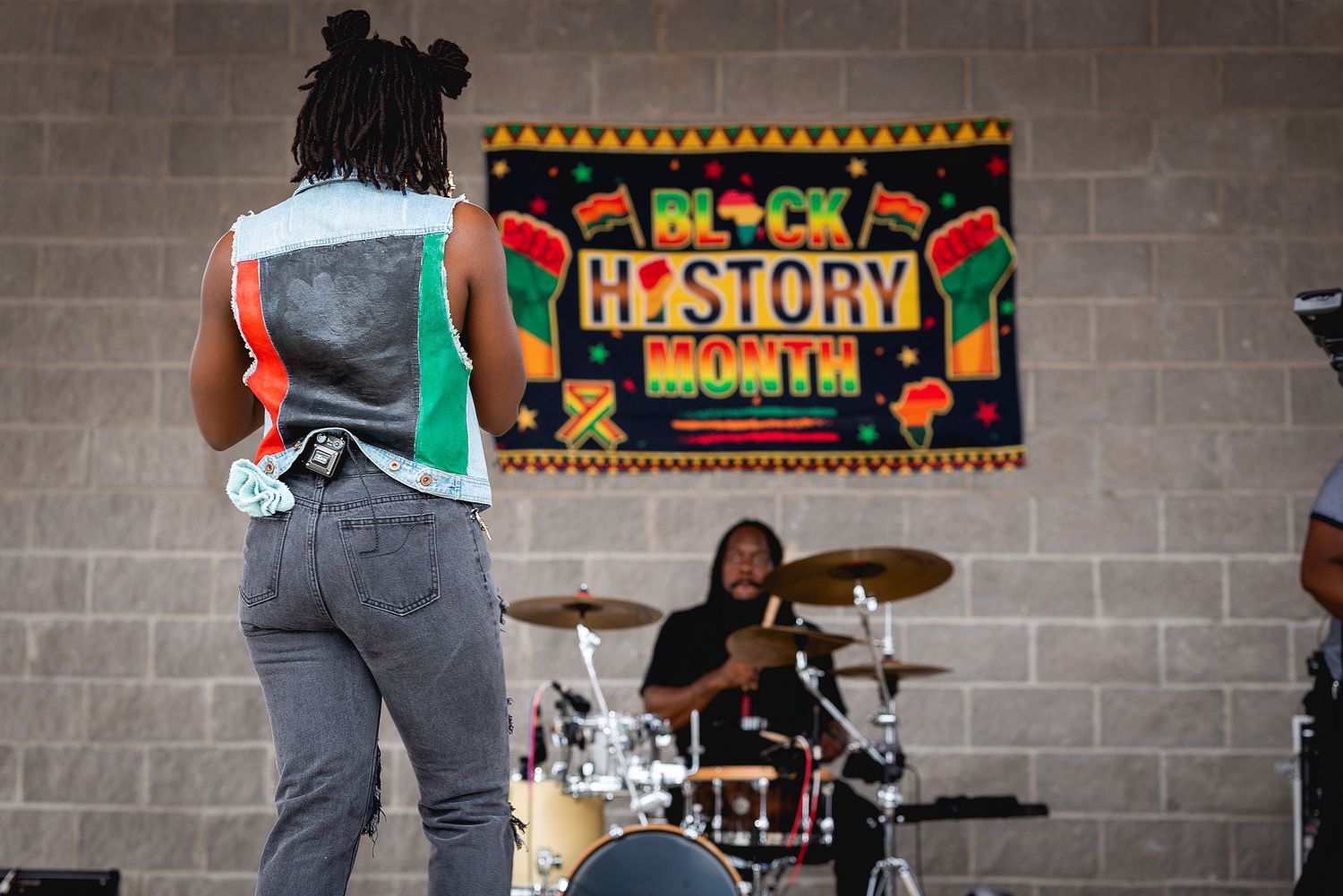 Musicians perform at a Black History Month event in front of a colorful banner against a brick wall backdrop.